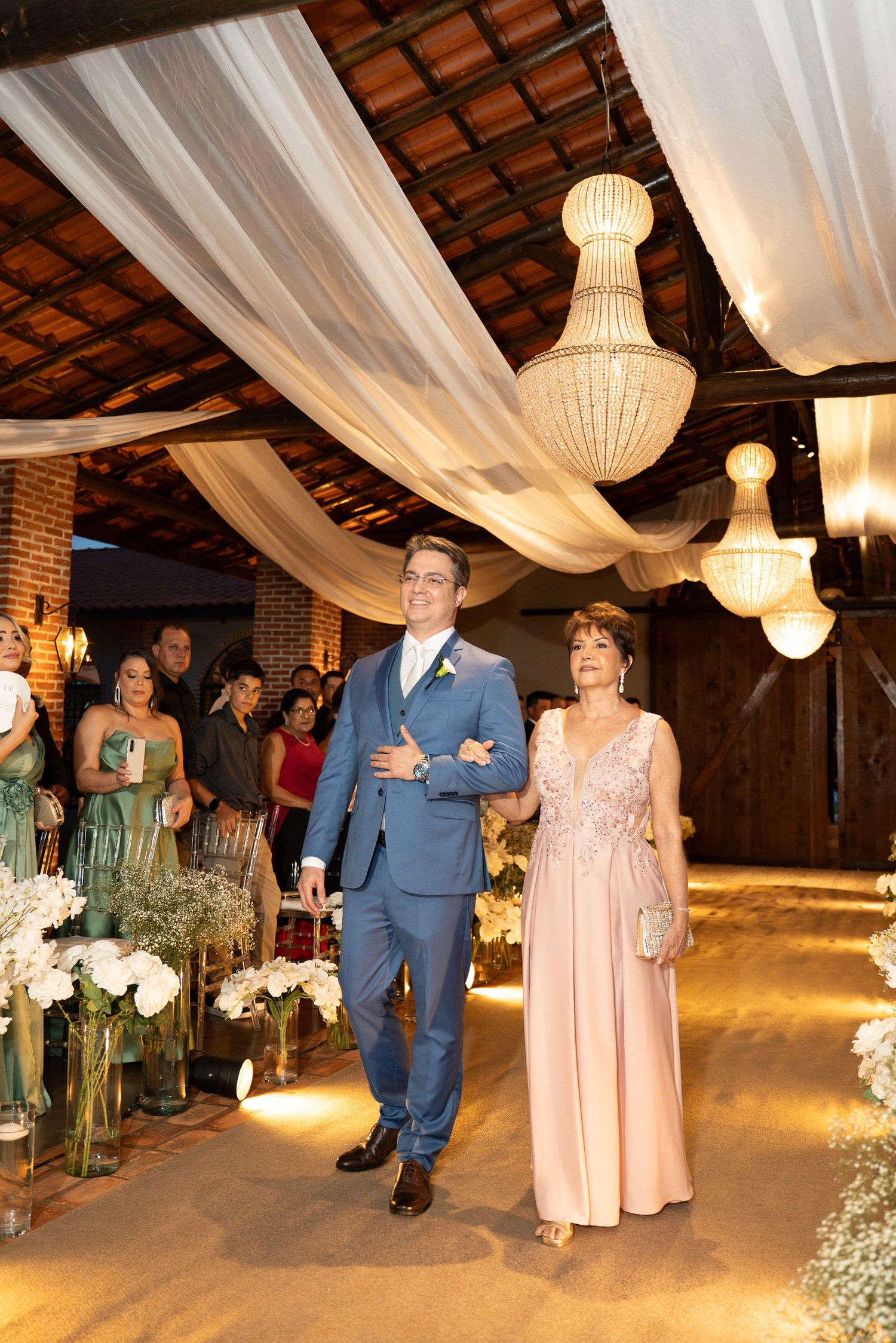 Groom in blue suit walks down aisle with mother in pink gown at elegant wedding venue with chandeliers.