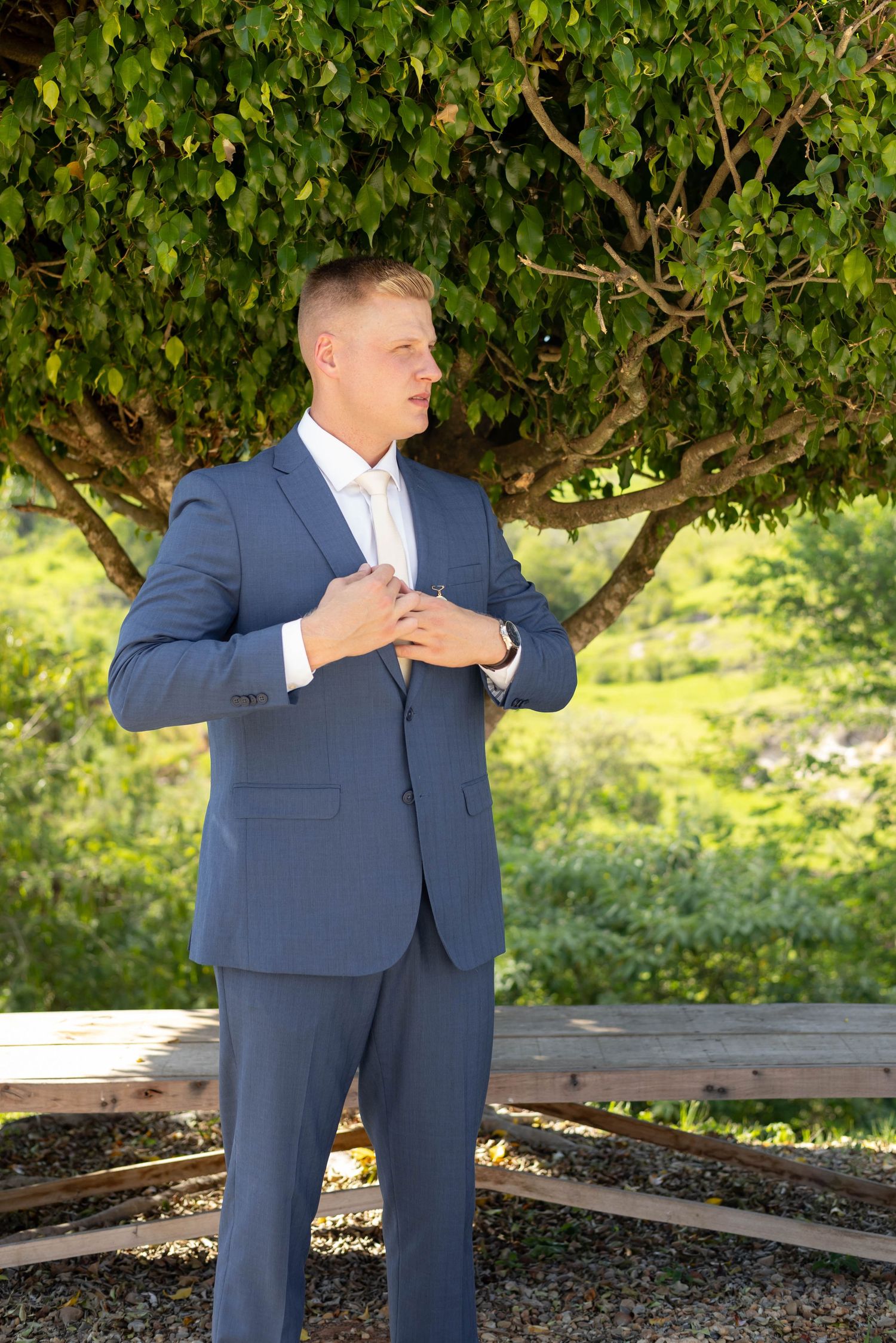 Young man in a navy blue suit adjusting his jacket outdoors under a green tree on a sunny day.