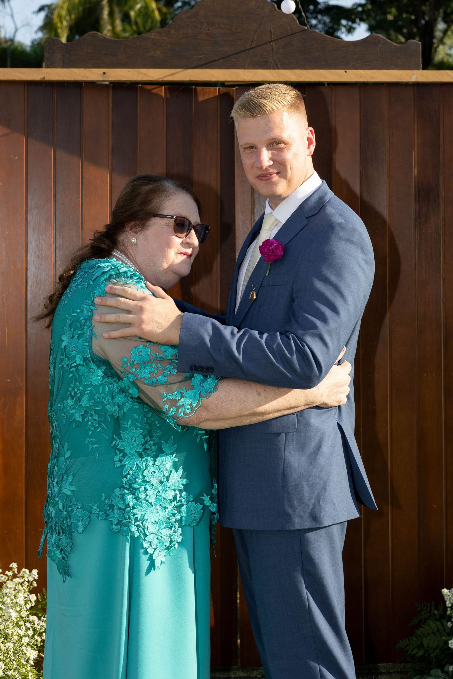 A groom in a navy suit with a pink boutonniere embraces a woman in a teal lace dress outdoors near a wooden fence.