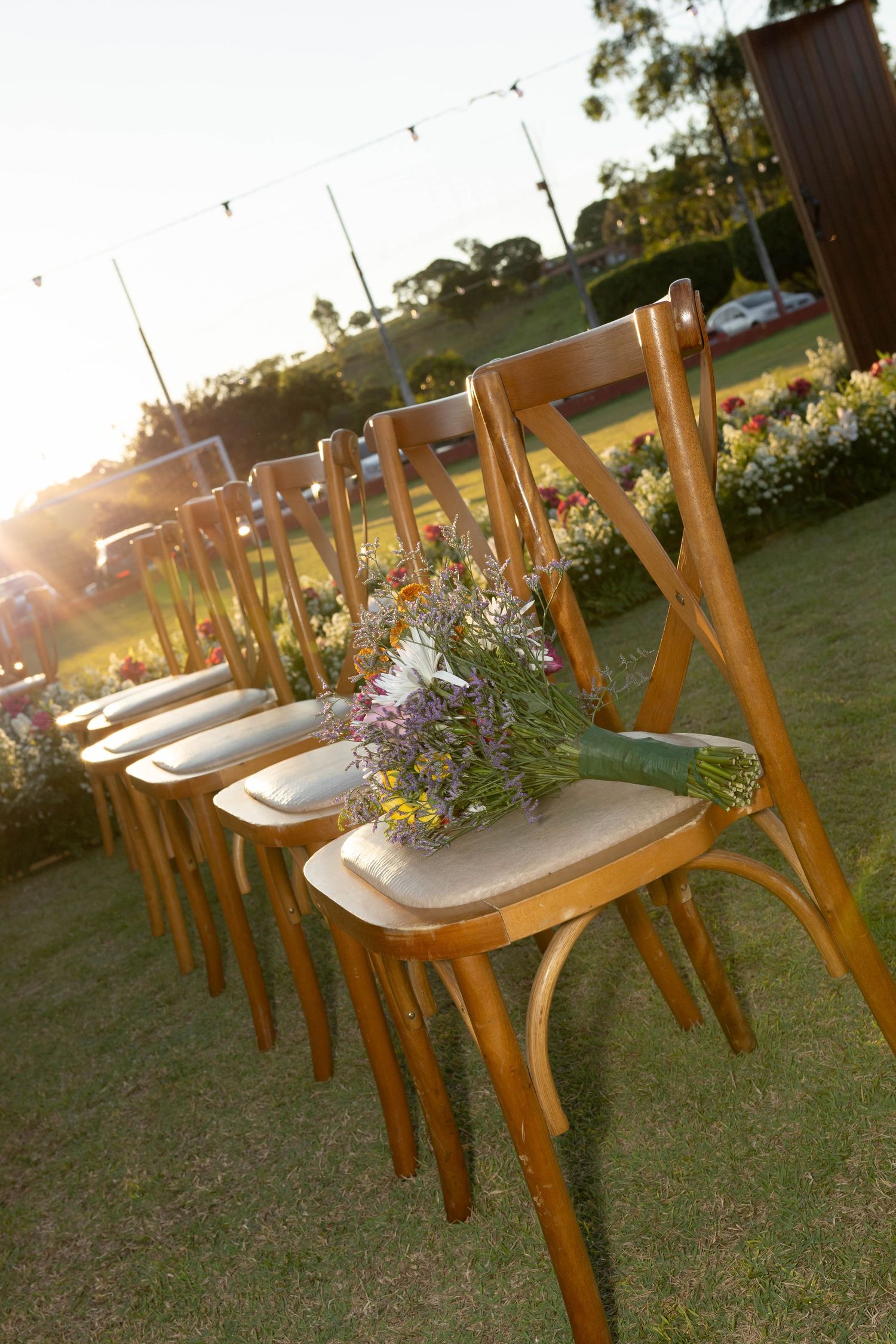 Wooden cross-back chairs arranged in rows on green grass with a wildflower bouquet, set for an outdoor wedding ceremony at sunset.