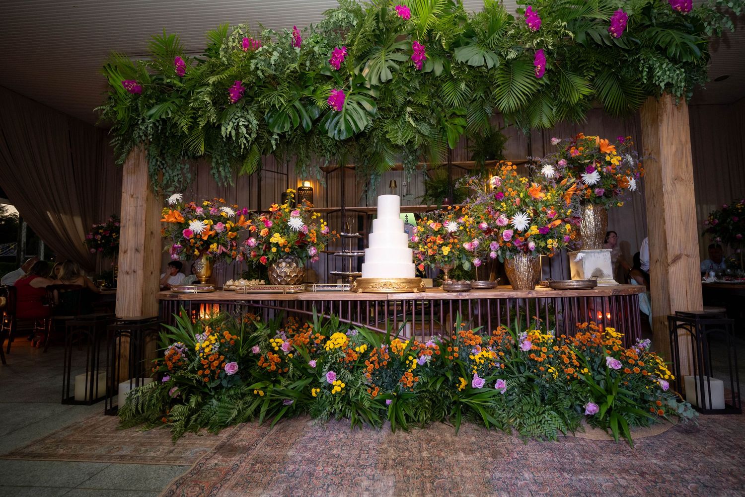 Lush tropical wedding cake table adorned with vibrant flowers, greenery arch, and a white tiered cake centerpiece.