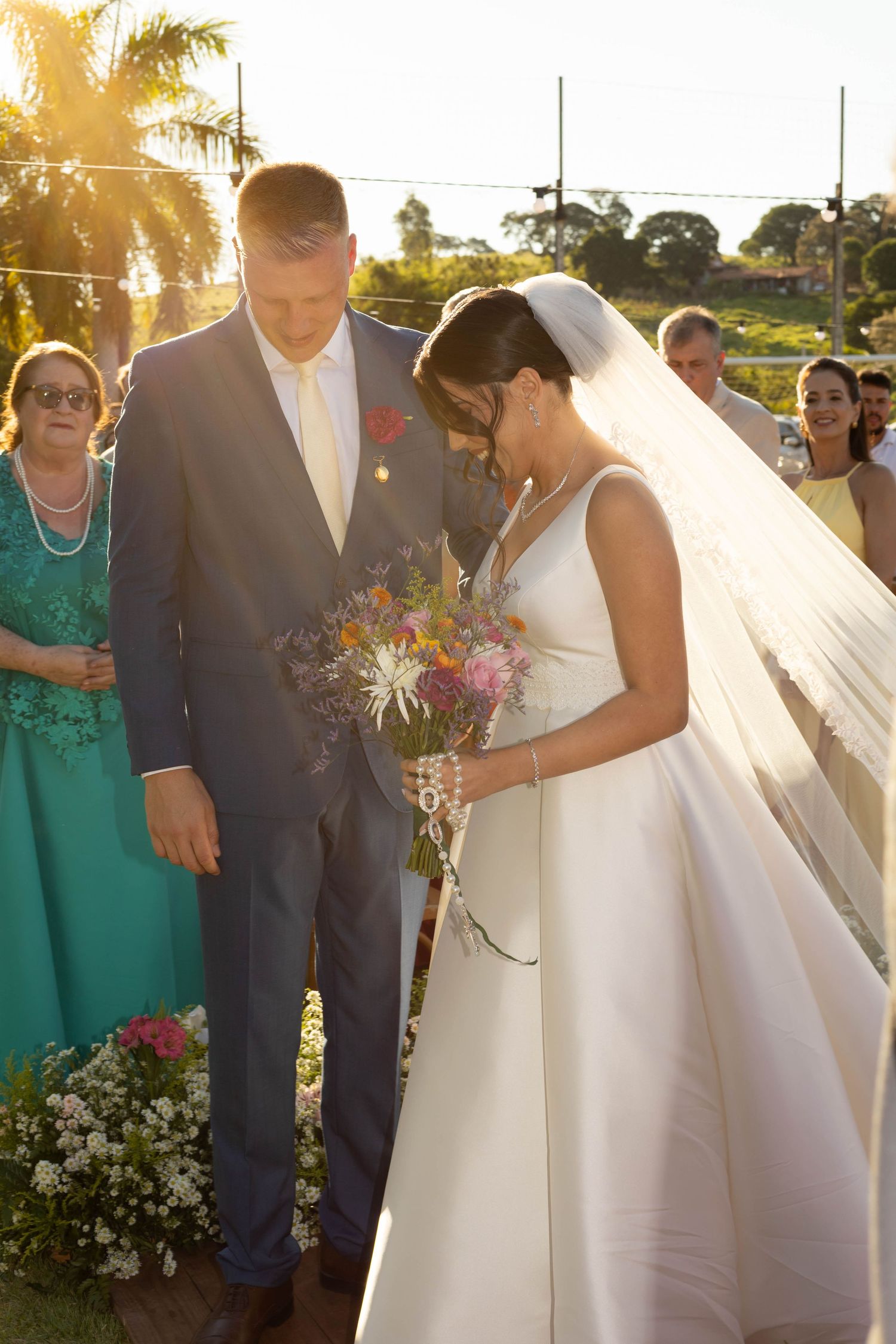 Bride and groom bow heads during outdoor wedding ceremony at golden hour, bride holding colorful bouquet in white gown.