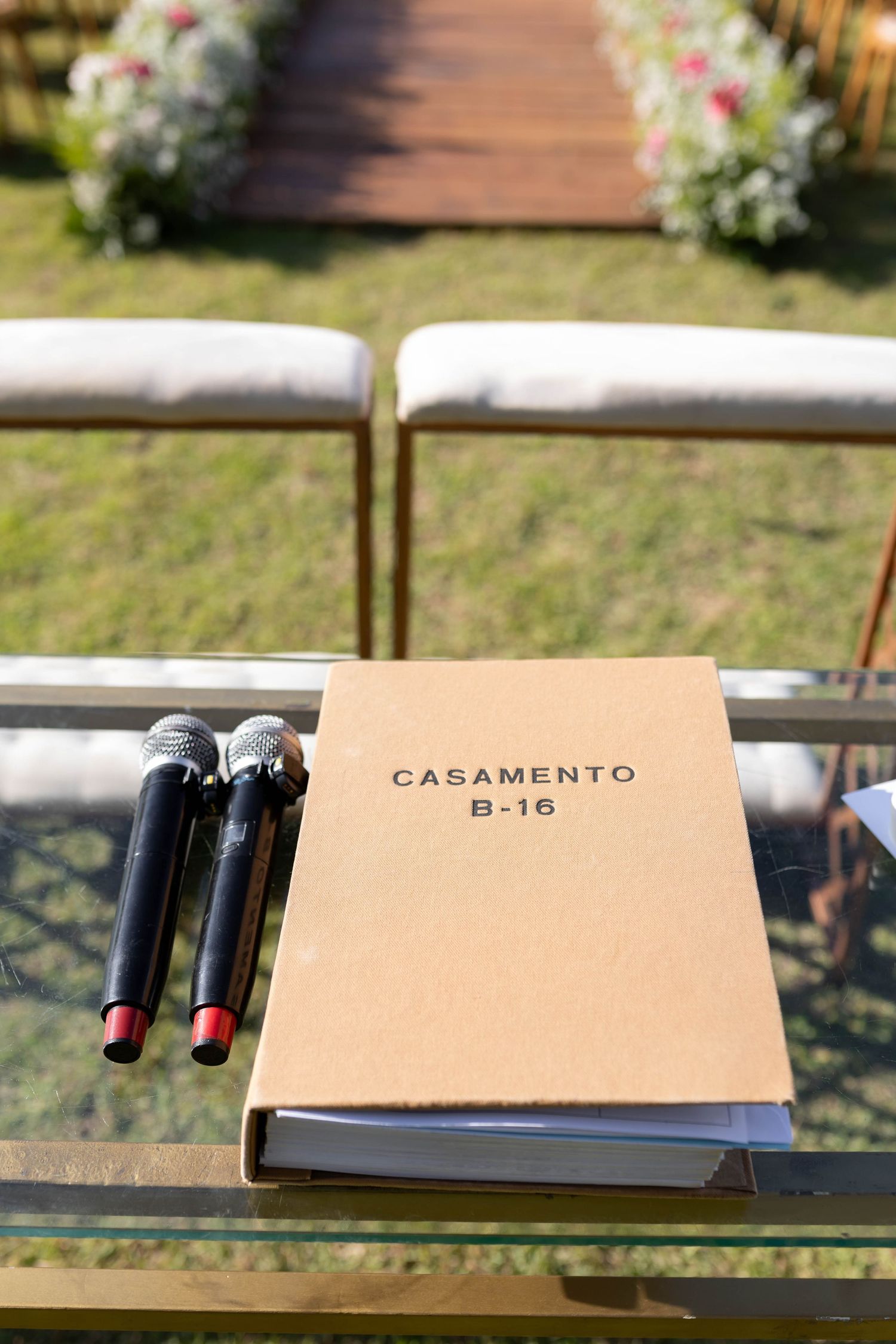 Outdoor wedding ceremony setup with microphones and a kraft paper folder labeled Casamento B-16 on a glass table.