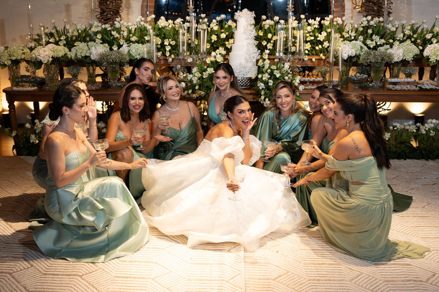 Bride in white gown sits on floor laughing with bridesmaids in sage green dresses at elegant wedding reception.