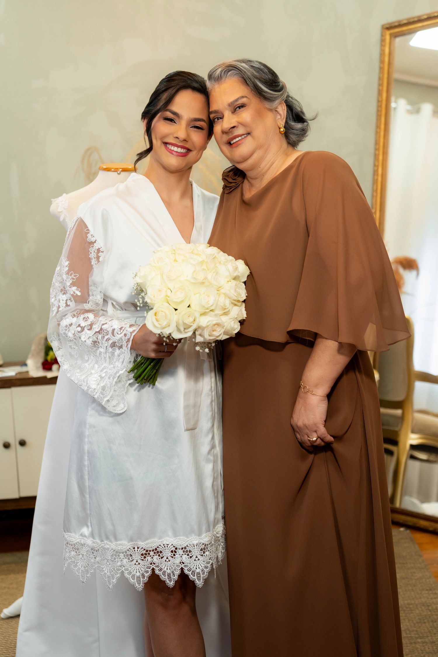 Bride in white lace robe holding bouquet of white roses poses with older woman in brown dress.