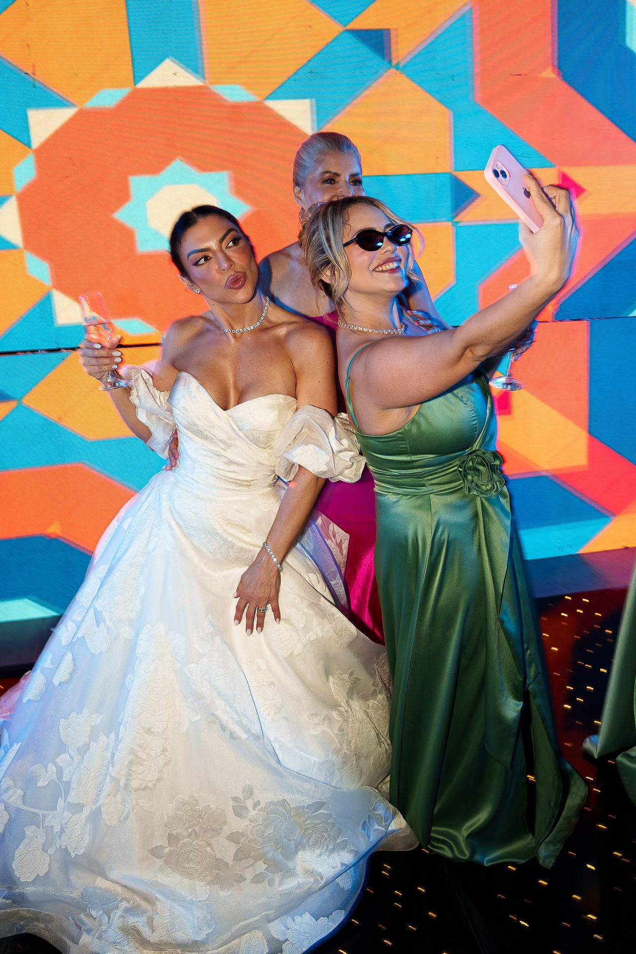 Bride in white gown takes selfie with friend in green satin dress and sunglasses at colorful wedding reception.