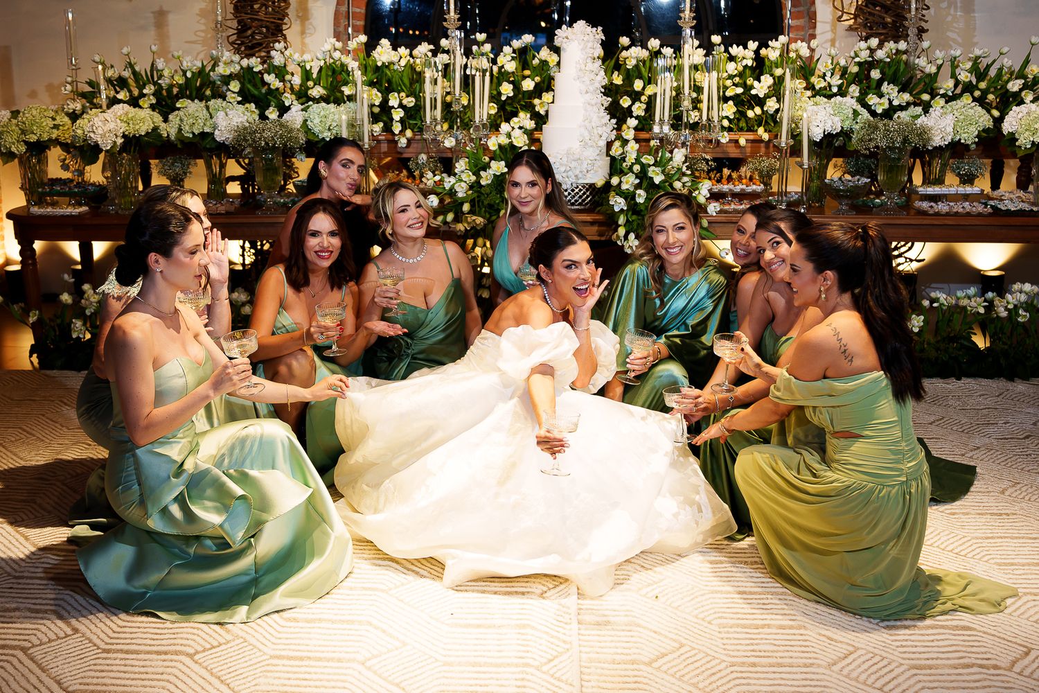 Bride in white gown sits on floor surrounded by bridesmaids in green dresses, celebrating at elegant wedding reception.
