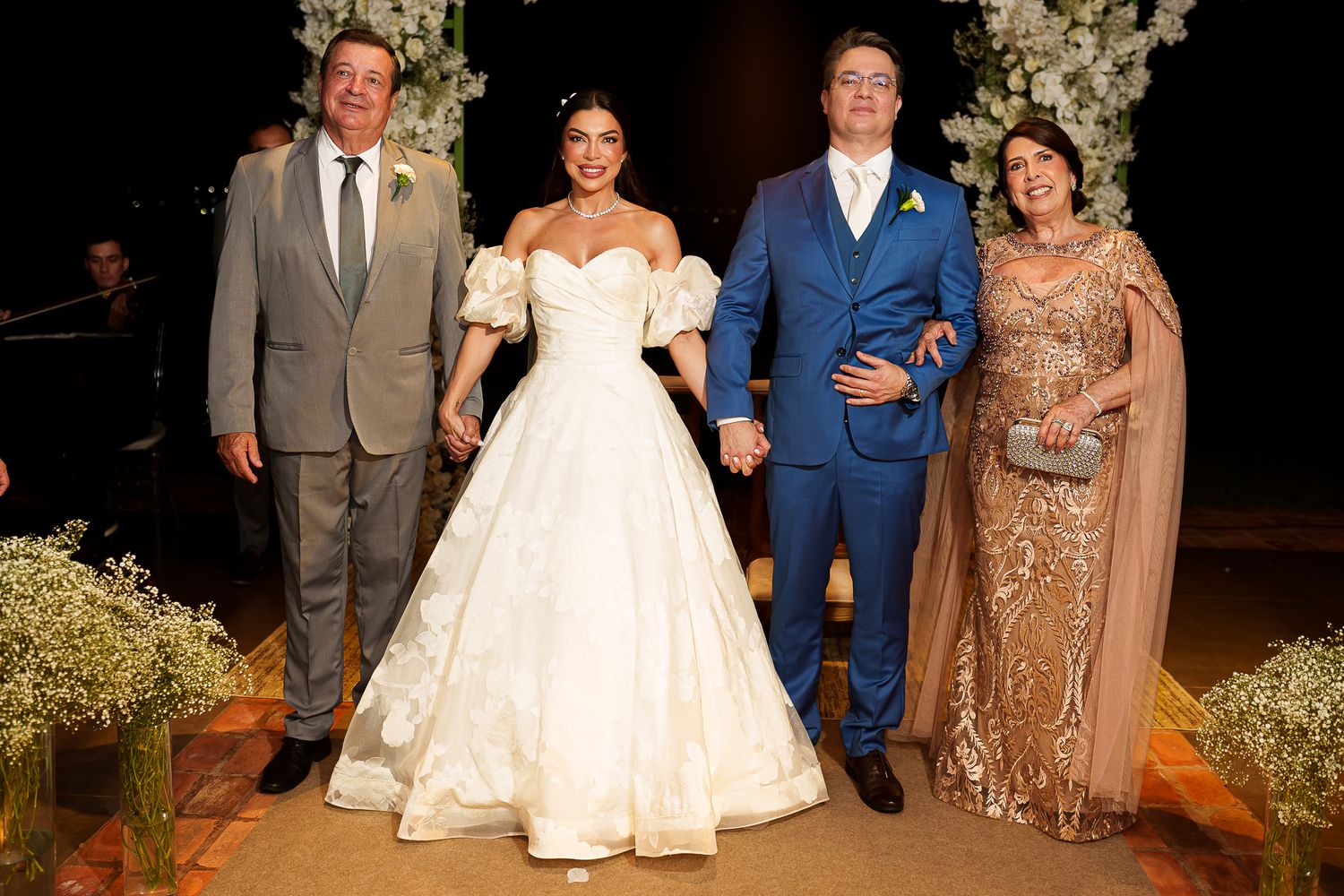 Bride in white ball gown and groom in blue suit pose with family at elegant floral wedding ceremony.