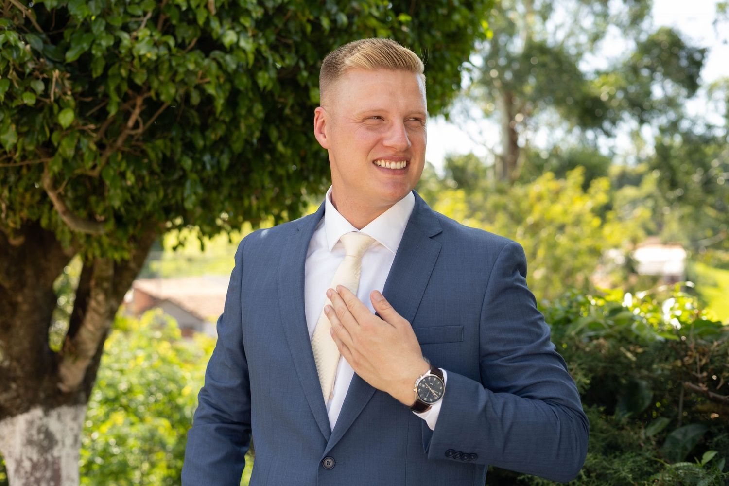 Smiling man in blue suit and gold tie adjusting his jacket outdoors with green trees in background.