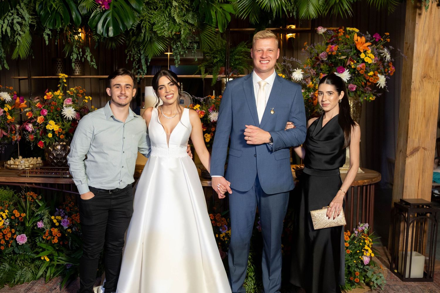 Wedding party of four posing before floral backdrop, bride in white gown with groom in blue suit flanked by guests.