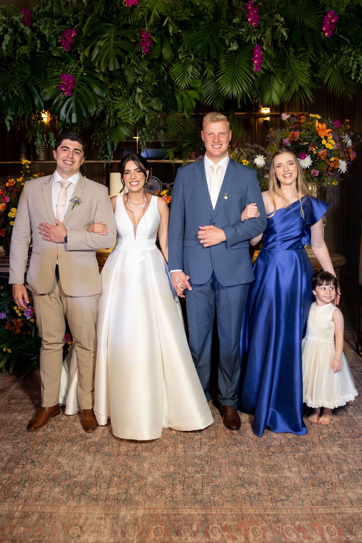 Wedding party posing in front of lush floral backdrop, bride in white gown with groom and guests in blue and beige formal attire.