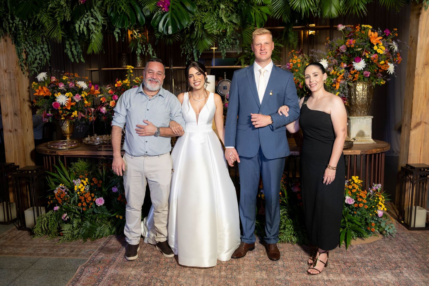 Bride in white gown and groom in blue suit pose with two guests amid lush floral decor at elegant wedding reception.