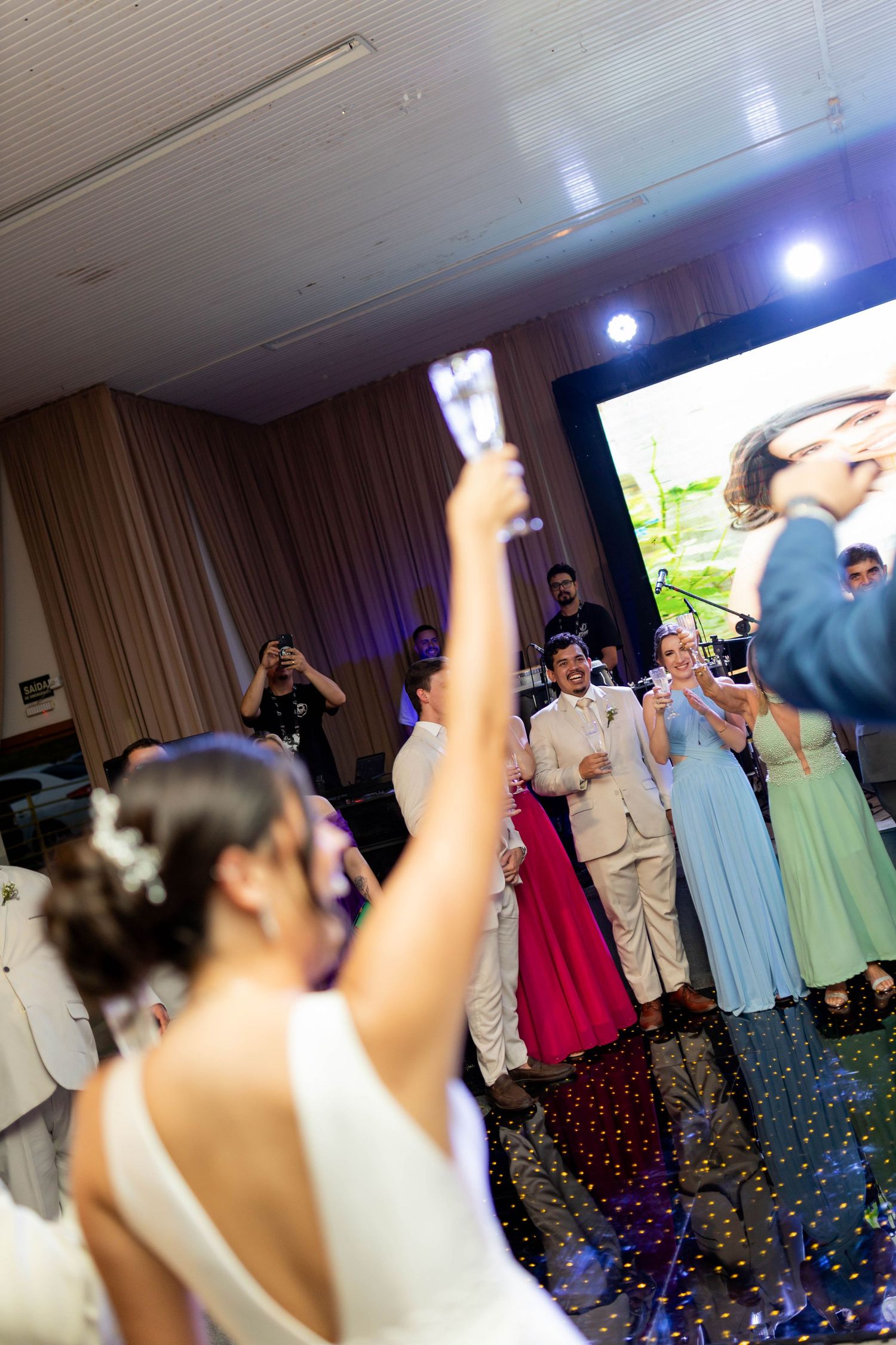Bride raises champagne glass at wedding reception with guests celebrating in background near large screen.