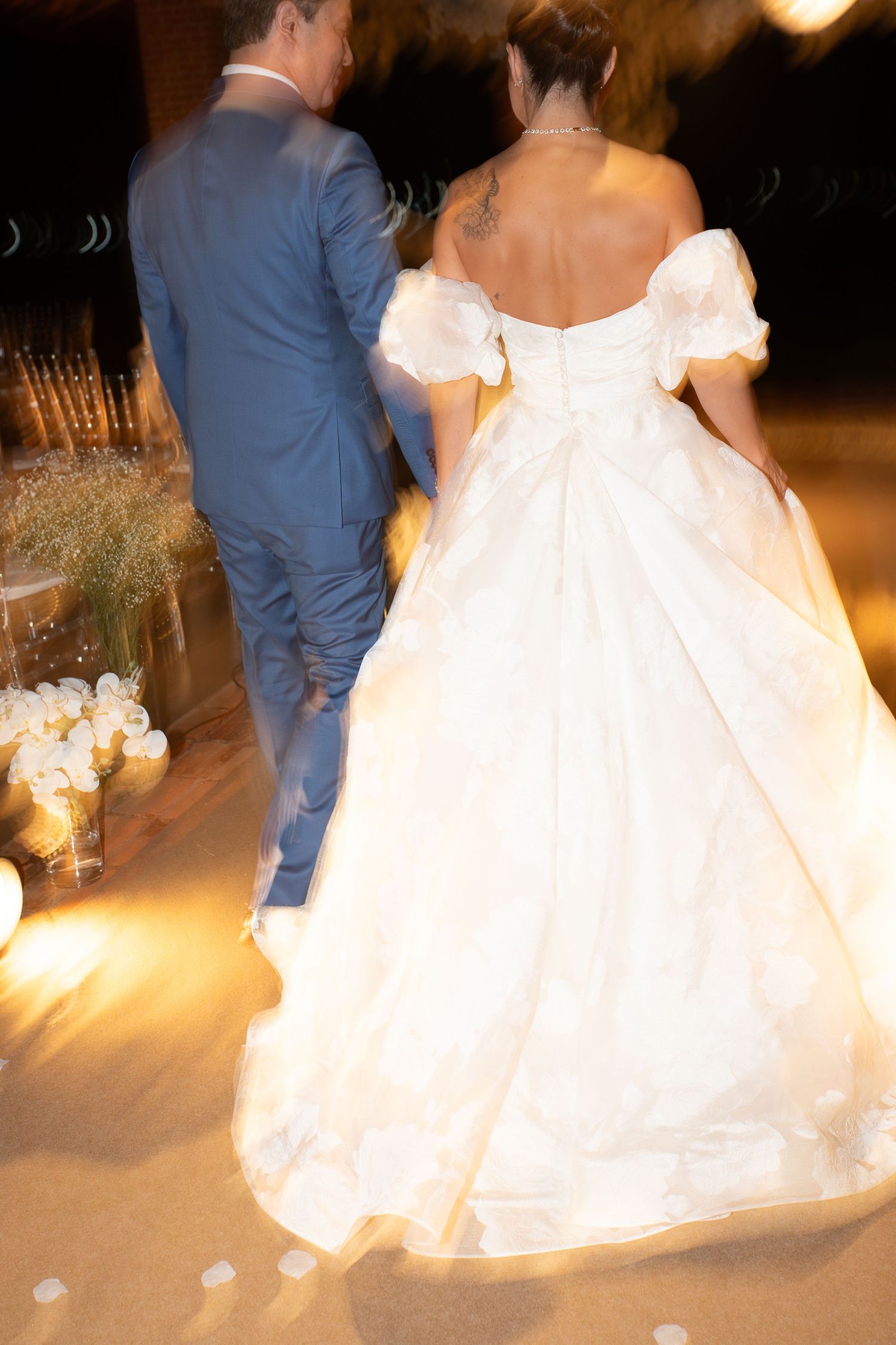 Bride in white off-shoulder ballgown walks with groom in blue suit at elegant candlelit wedding reception.