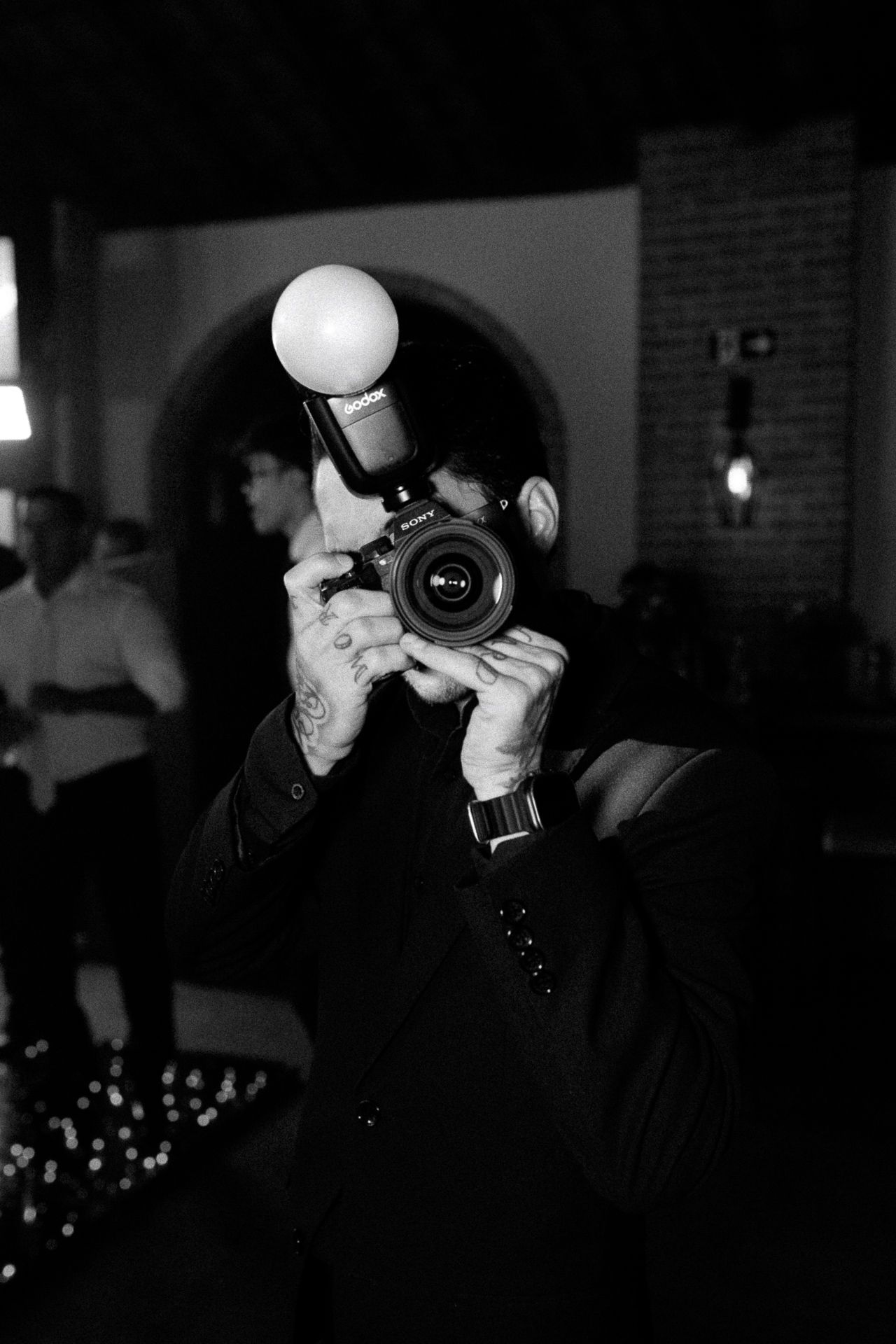 Black and white photo of a tattooed photographer holding a DSLR camera with flash bulb at an indoor event.