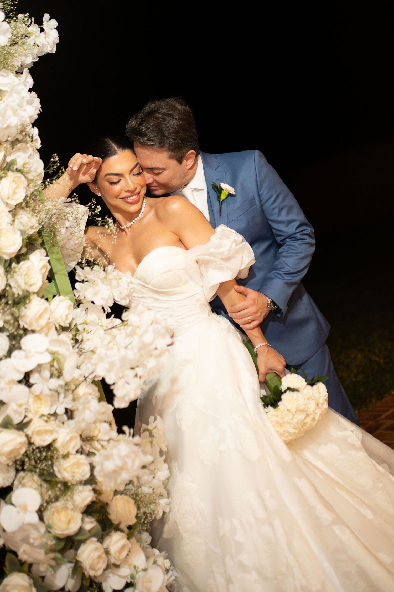 Bride in white off-shoulder gown and groom in blue suit share a romantic kiss beside white floral arrangements at night.