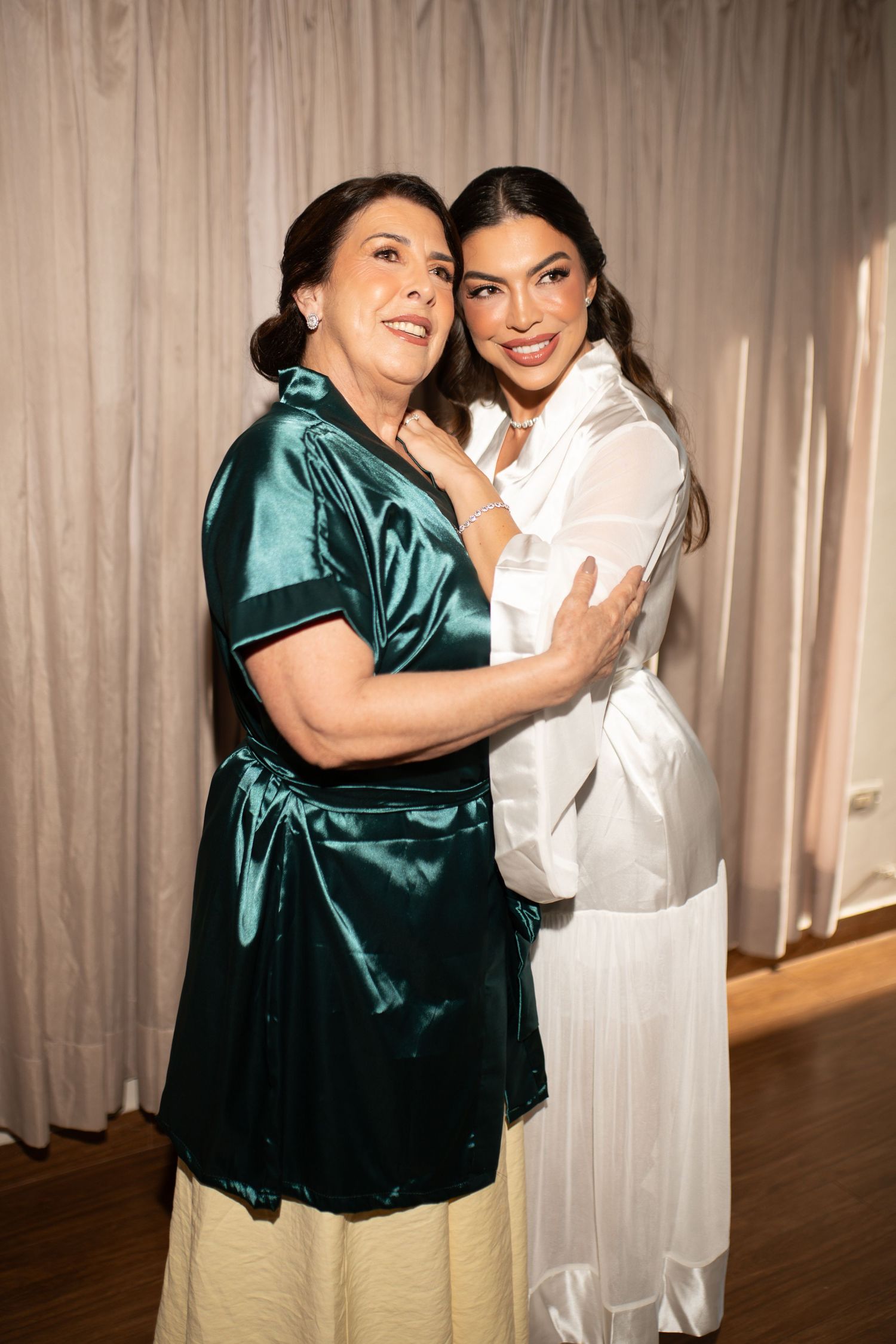 Two women in elegant satin outfits embracing and smiling warmly in a softly lit indoor setting.