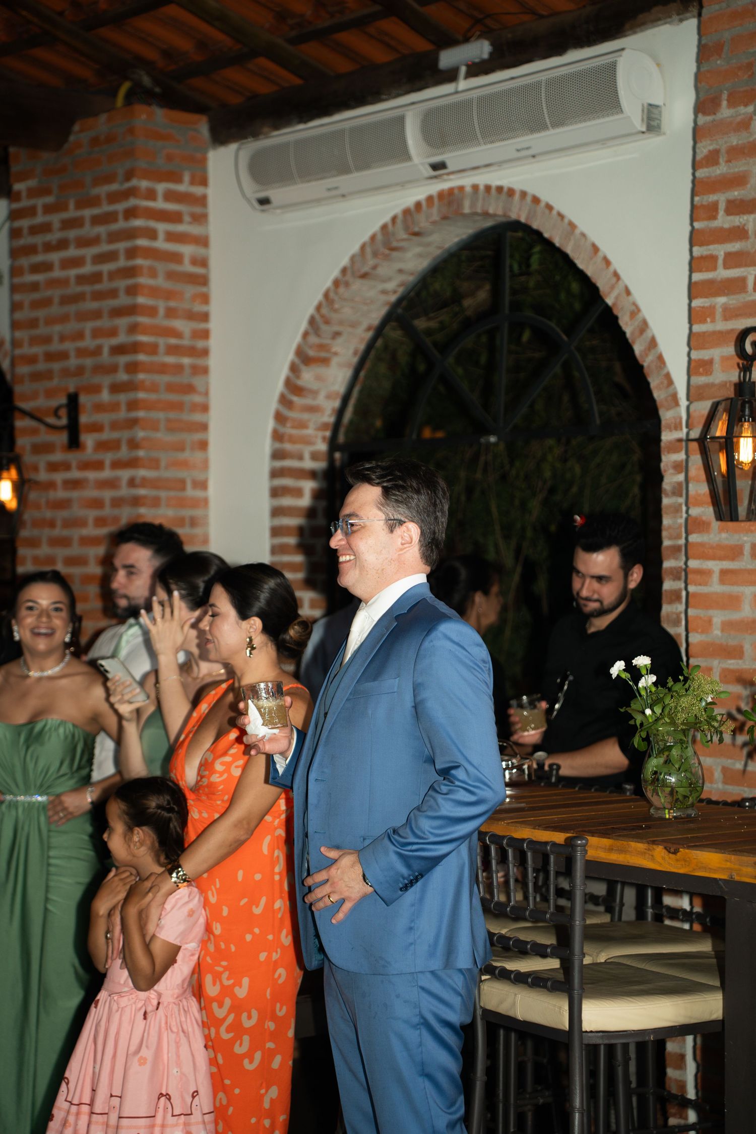 Guests in formal attire at an elegant wedding reception venue with brick arches, including a man in a blue suit.