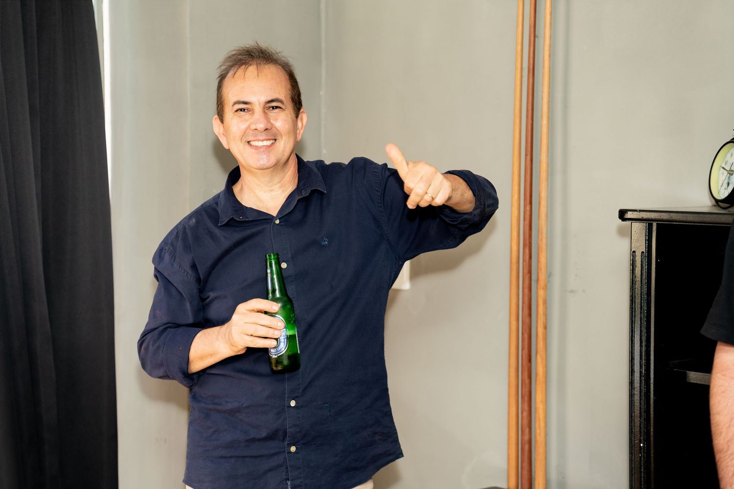 Smiling man holding a green beer bottle giving a thumbs up gesture indoors at a casual gathering.