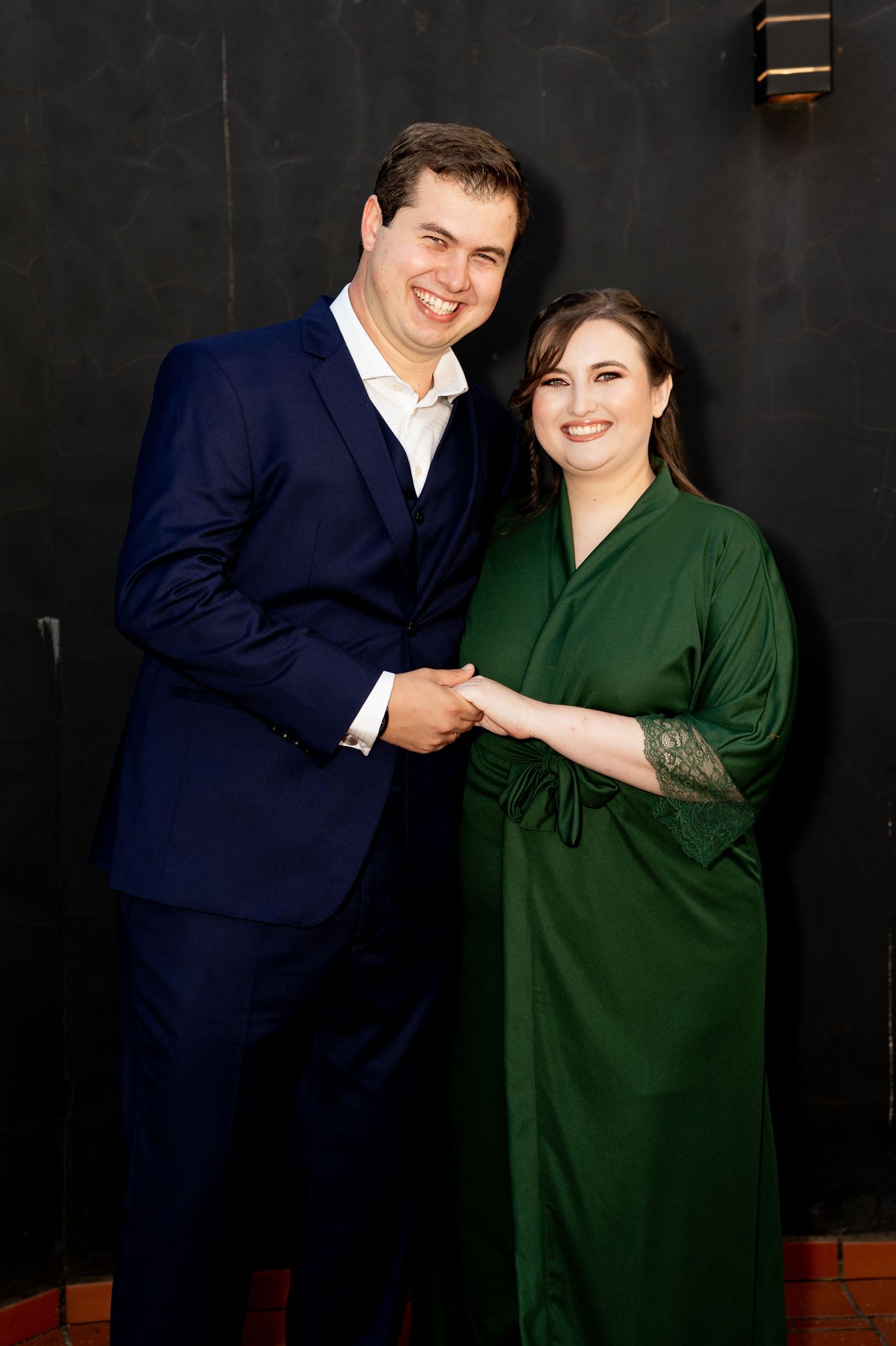 A smiling couple poses together, man in navy suit and woman in elegant green dress, holding hands against dark wall.