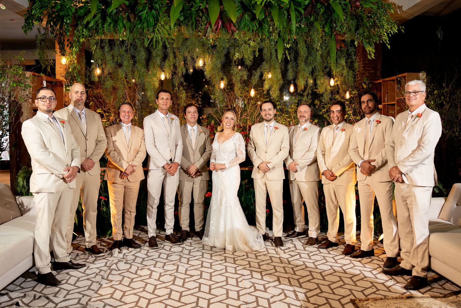 Bride in white gown poses with groomsmen in tan suits under lush greenery and string lights at elegant wedding venue.