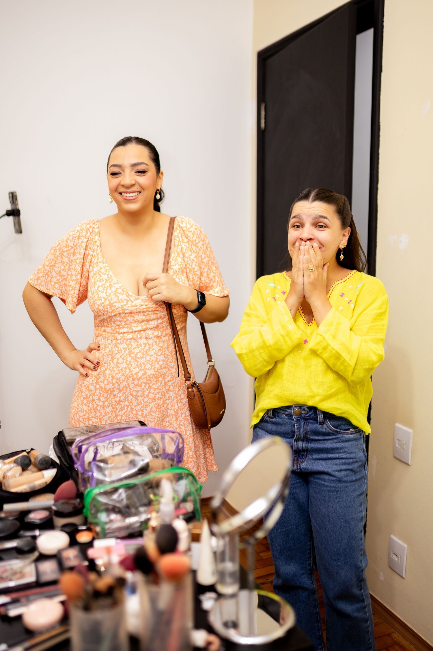 Two women smile near a makeup table with beauty products, one in an orange dress, the other in a yellow top and jeans.
