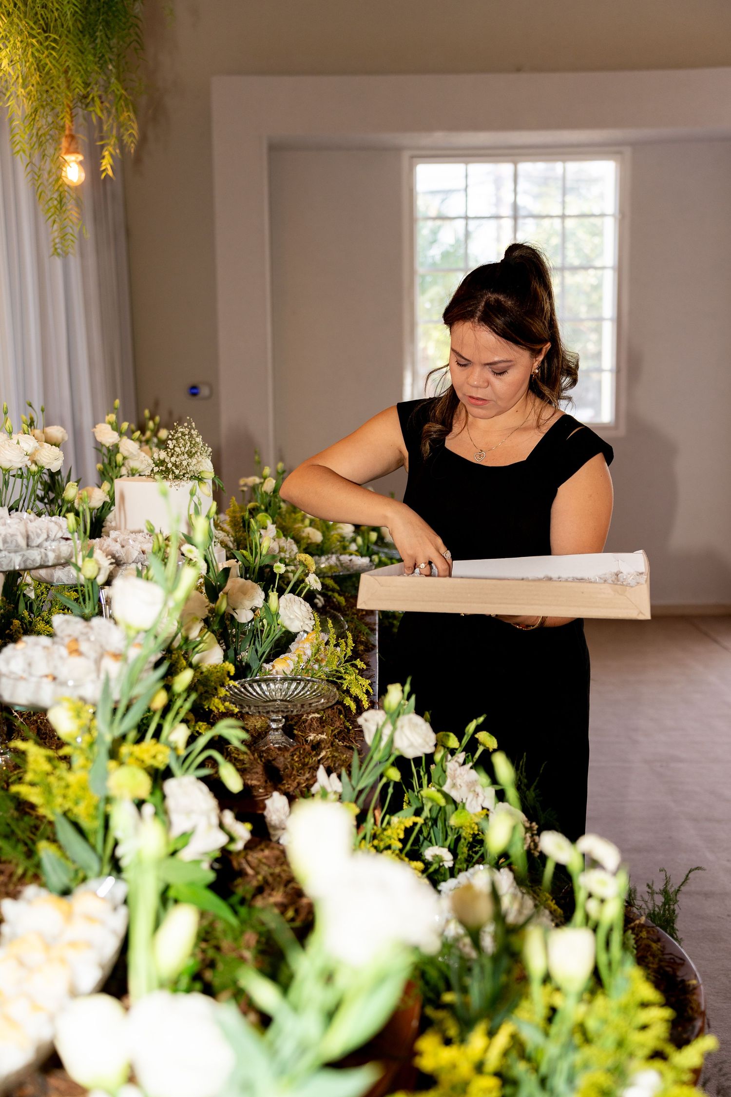 Woman in black dress arranging white floral centerpieces on decorated event table with roses and greenery.