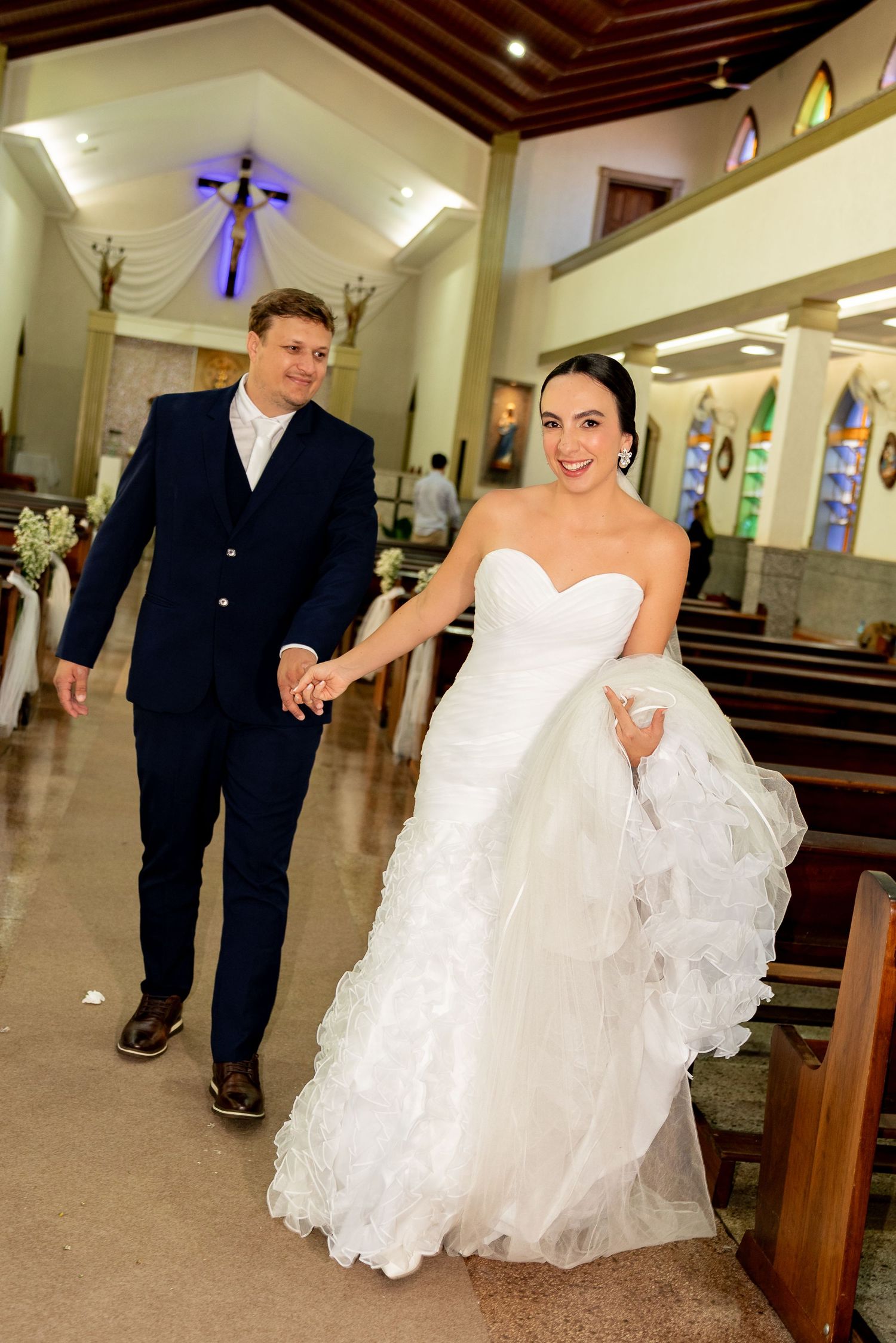 Bride and groom walking hand in hand down church aisle after wedding ceremony, bride in white ruffled gown.