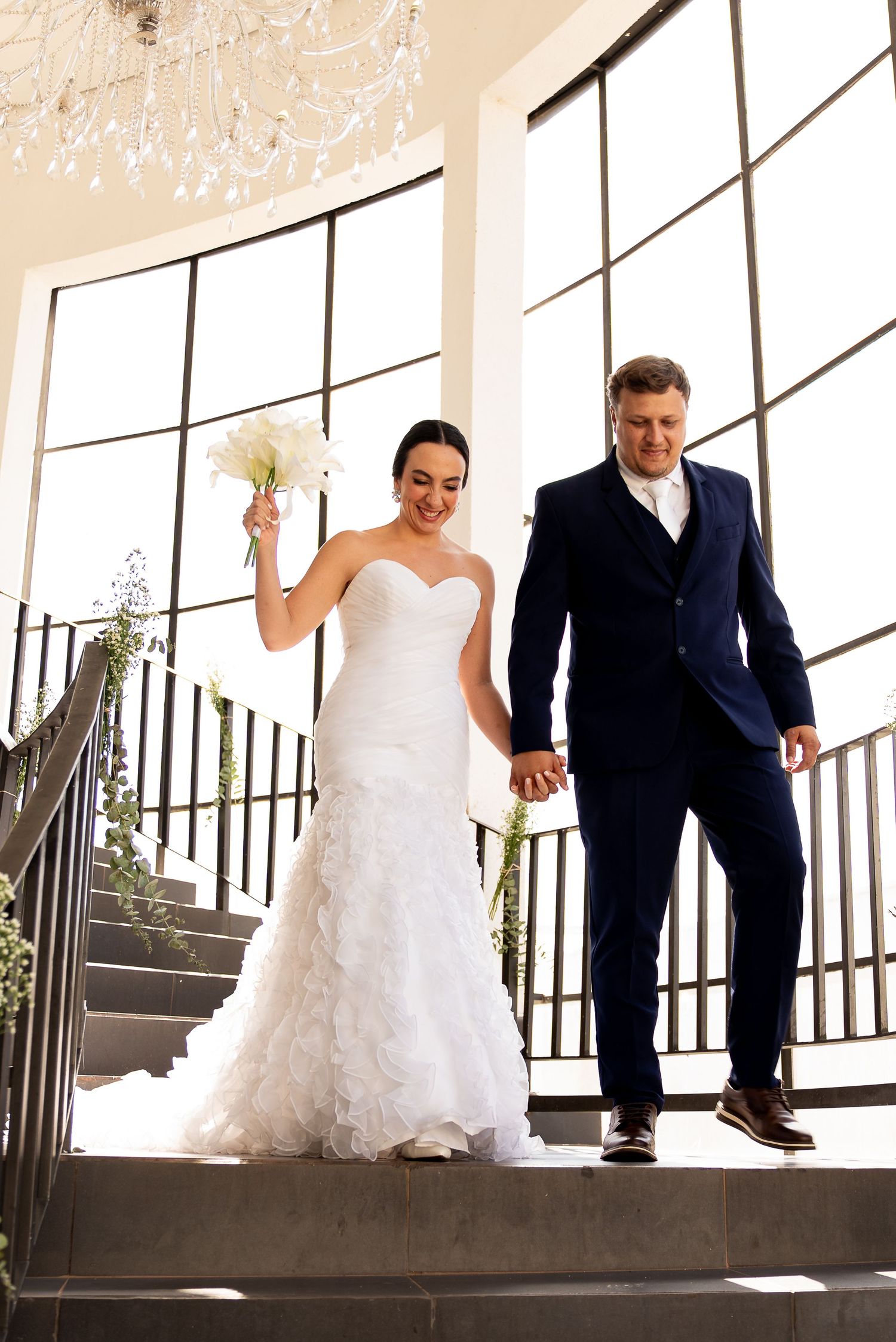 Bride in white ruffled gown and groom in navy suit descend grand staircase holding hands at elegant wedding venue.