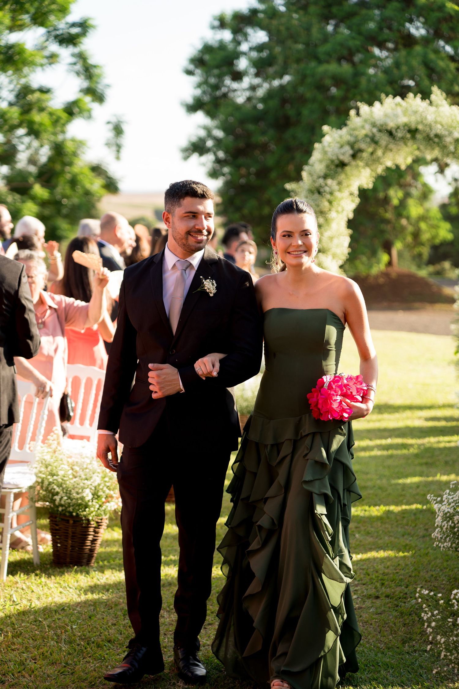 A smiling couple walks down an outdoor wedding aisle, man in dark suit, woman in green strapless gown holding pink bouquet.