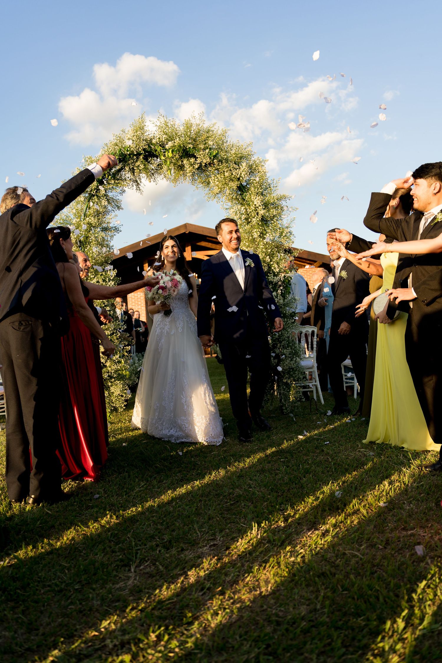 Newlyweds walk through floral arch as guests toss petals at outdoor wedding ceremony on sunny day.
