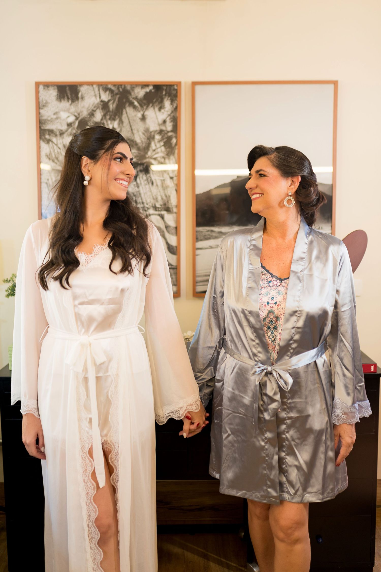 Bride and bridesmaid in satin robes holding hands and smiling at each other during wedding preparations.