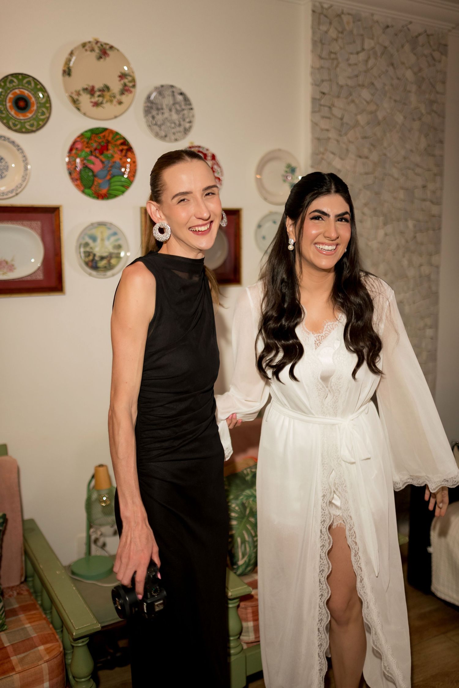 Two women smiling, one in black gown and one in white lace bridal robe, posing together indoors with decorative plates on wall.