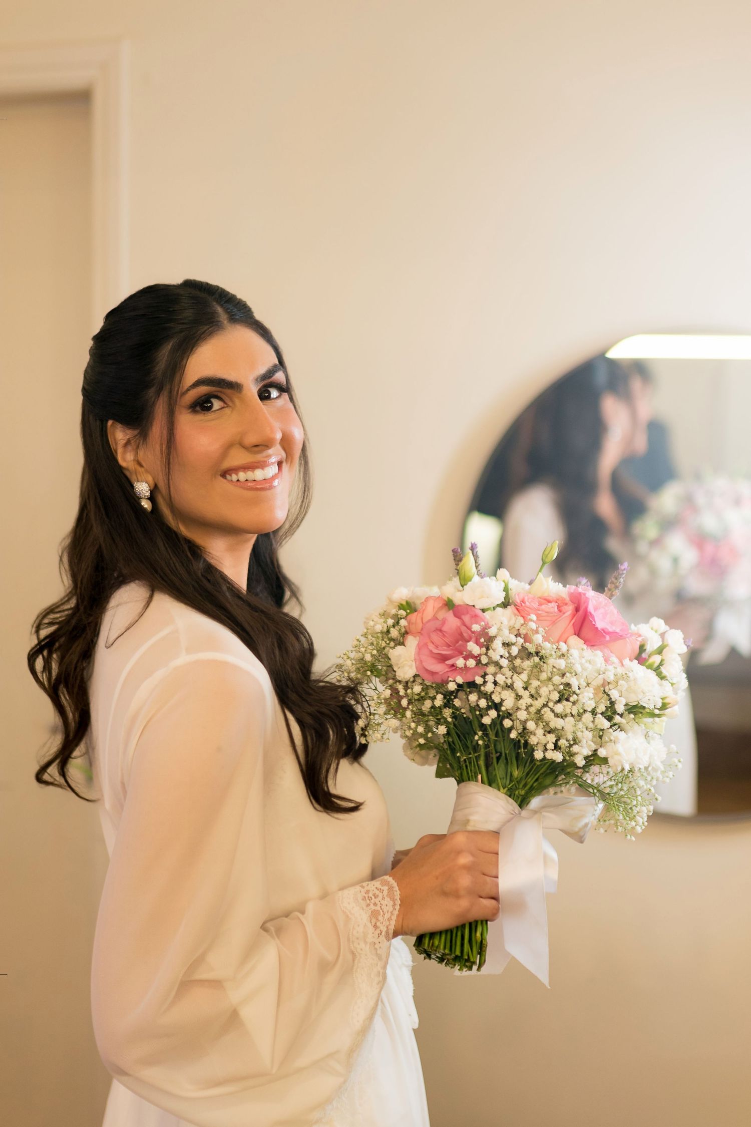 Smiling bride in white robe holds pink rose bouquet with baby's breath, standing before a mirror in a bridal suite.
