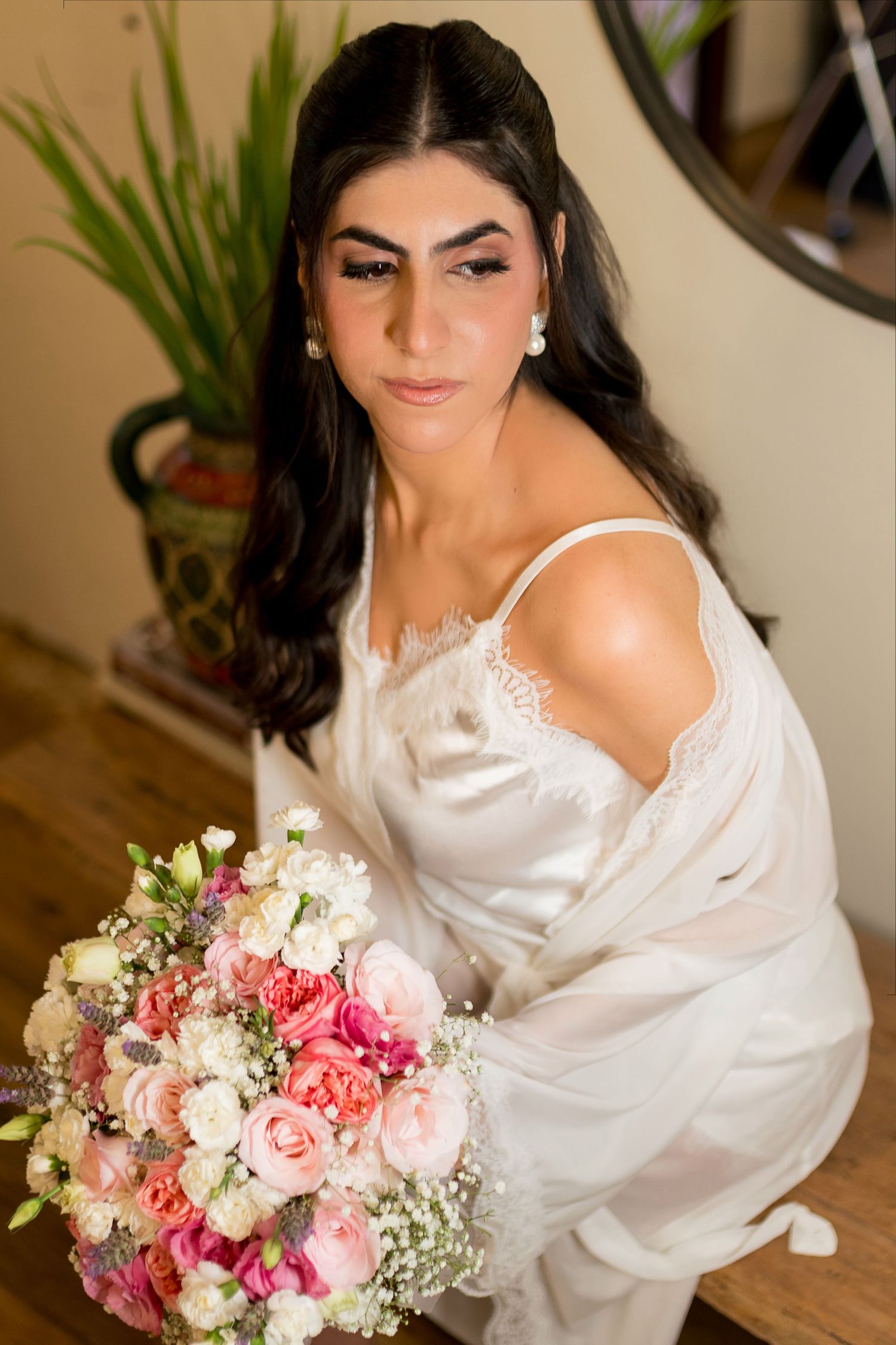 Bride in white lace robe holding large pink rose bouquet with dramatic bridal makeup.