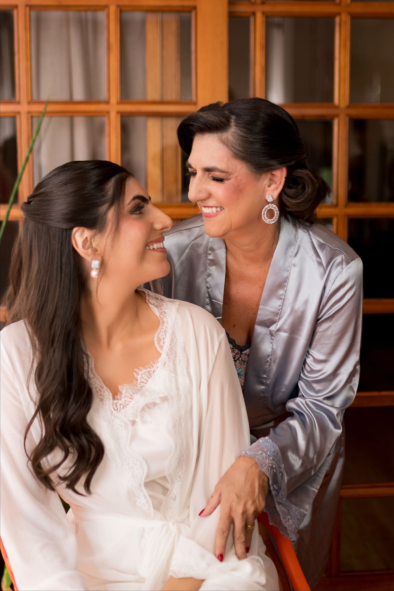 Two smiling women in satin robes share a joyful moment during bridal preparations, wearing elegant drop earrings.