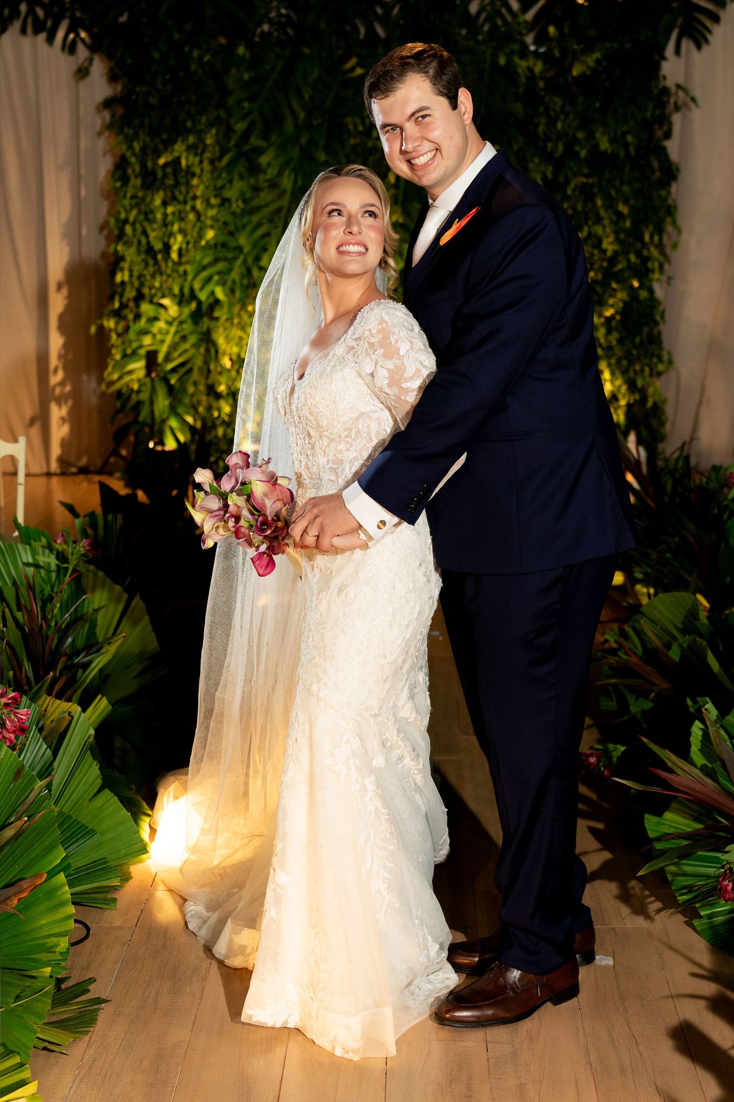 A bride in lace gown with veil holding bouquet poses with groom in navy suit amid tropical greenery.