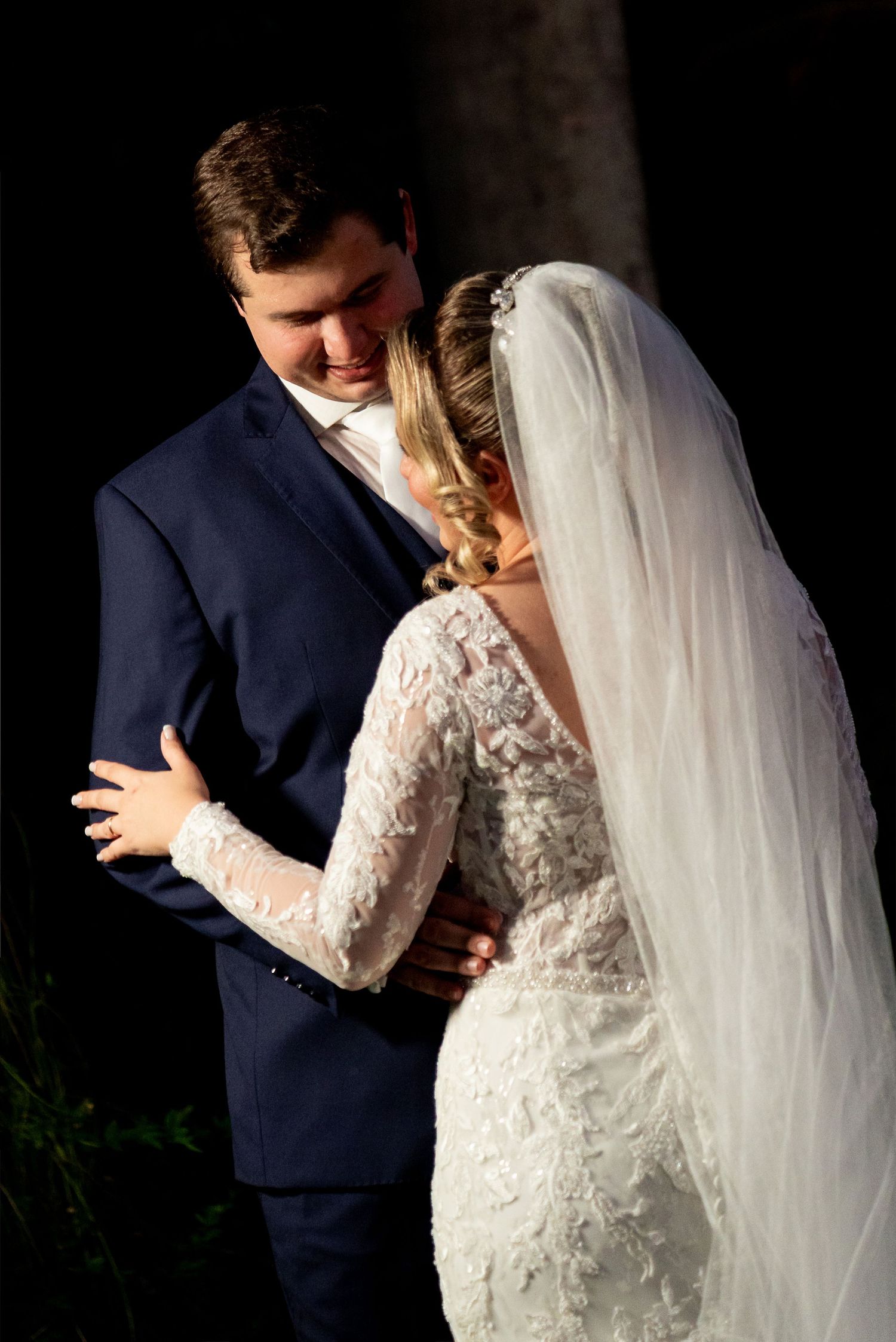 Bride in lace gown and veil embraces groom in navy suit during romantic outdoor wedding ceremony.