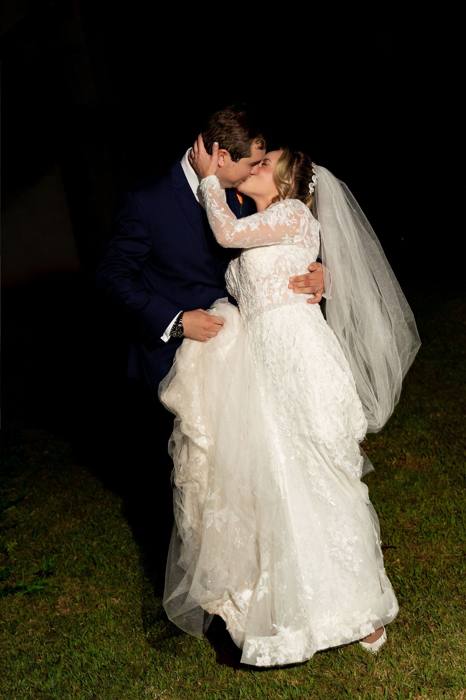 Bride and groom sharing a romantic kiss outdoors at night, bride wearing lace wedding gown with veil.