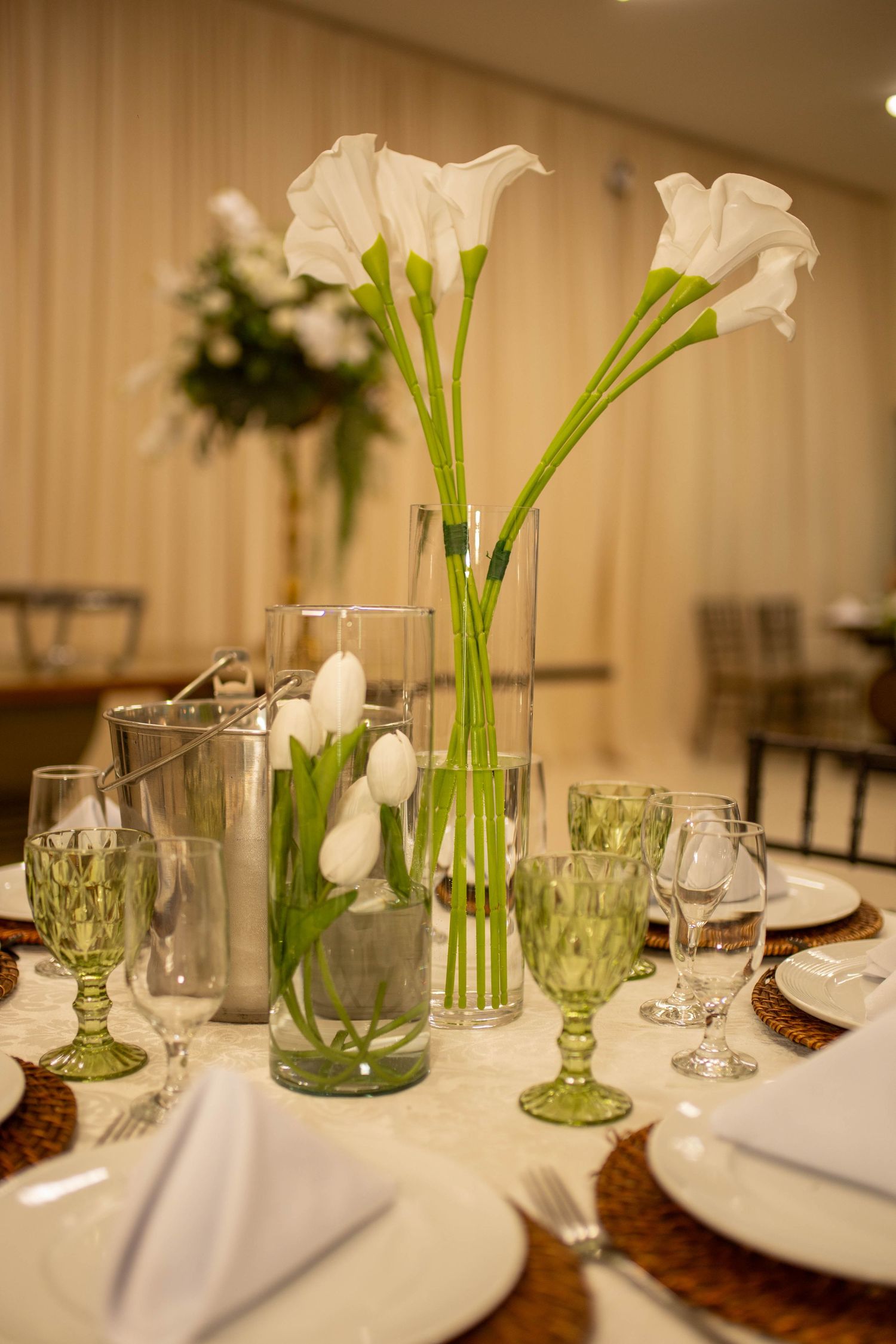 Elegant wedding table setting with white calla lilies in glass vases, green goblets, white plates, and wicker chargers.