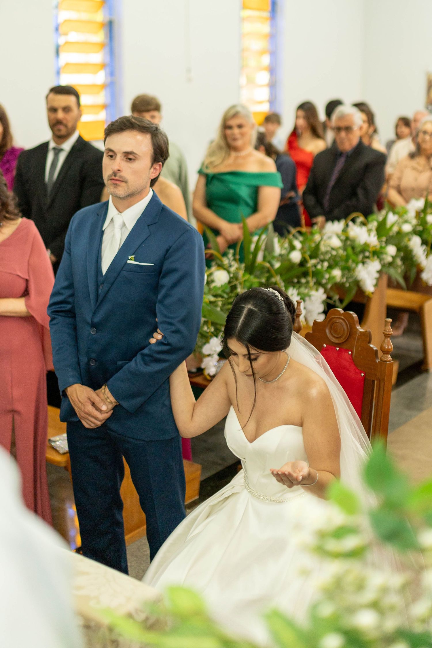 Bride and groom kneeling at church altar during wedding ceremony, guests seated in pews with floral decorations.