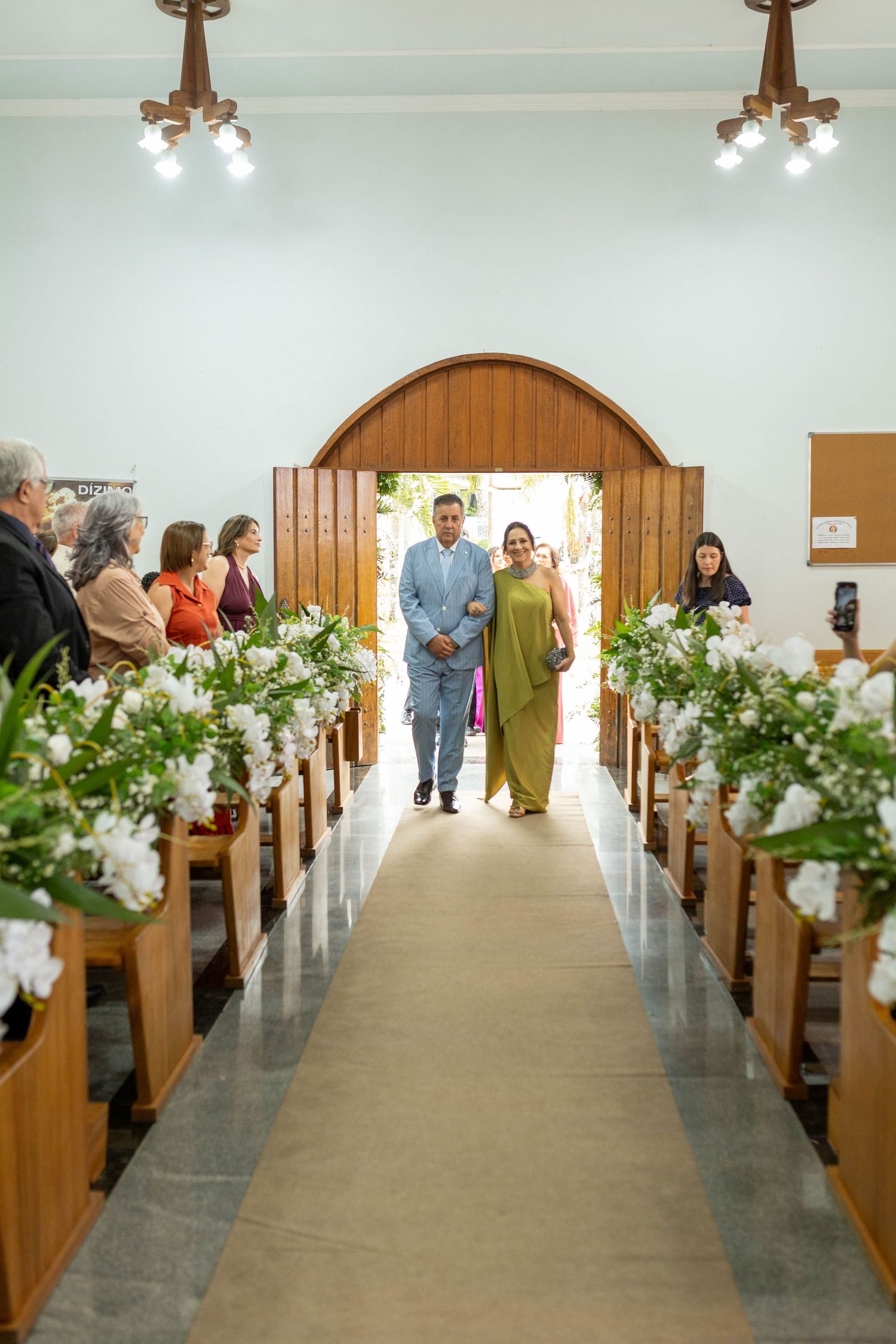 Couple walks down a flower-adorned church aisle during a wedding ceremony, with guests seated on wooden pews.
