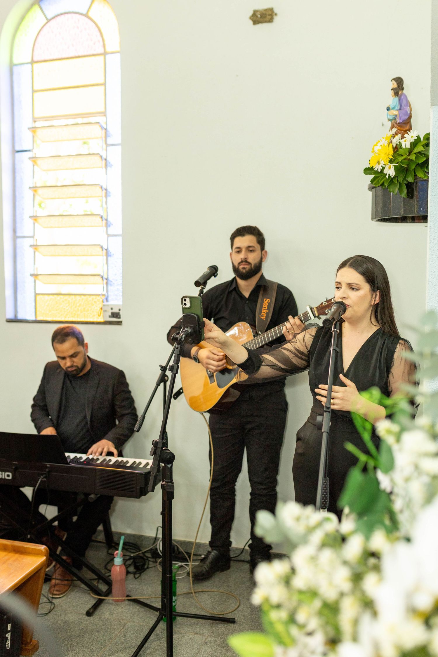 Three musicians perform in a church, with a keyboardist, guitarist, and female vocalist dressed in black.