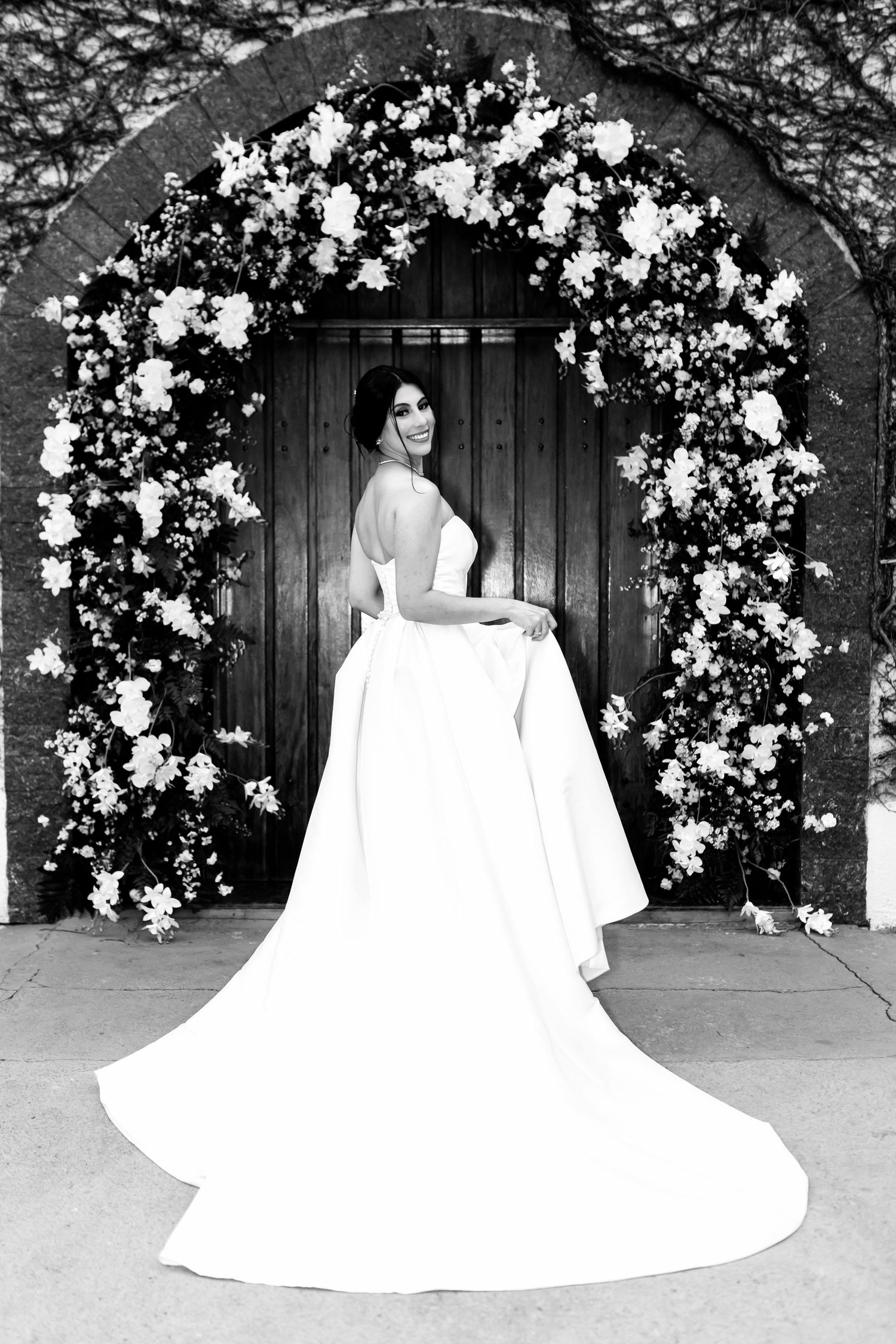 Black and white photo of a bride in a elegant gown with long train standing under a floral arch doorway.