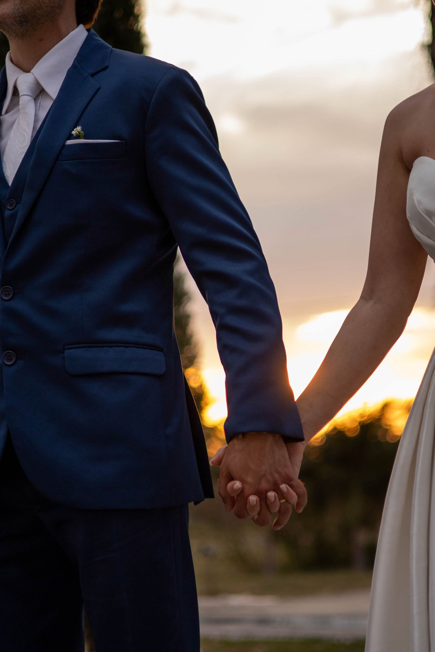 Bride and groom holding hands at sunset, groom in navy blue suit with white pocket square and boutonniere.