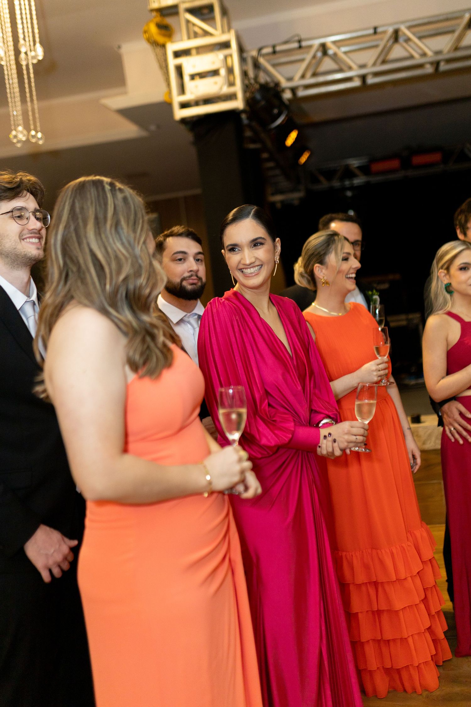 Elegantly dressed guests in vibrant pink and orange gowns socializing at a formal event, holding champagne glasses.