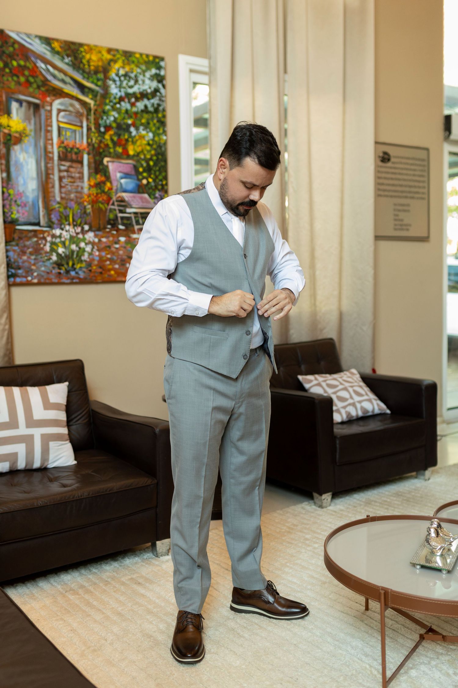 Man in gray vest and trousers buttoning his suit in an elegant room with colorful artwork.