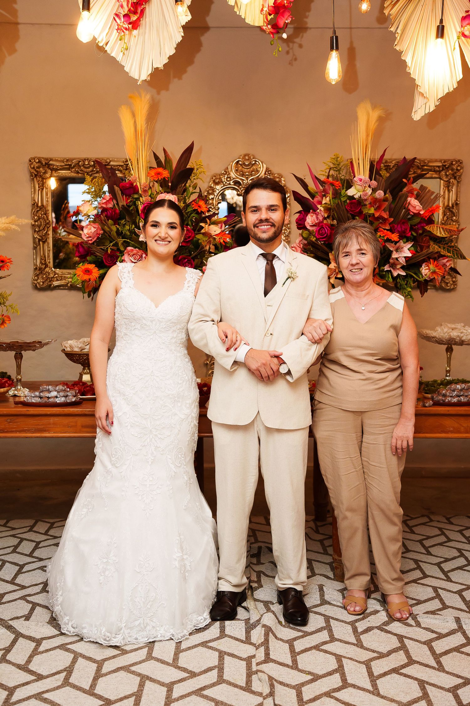 Bride in white gown, groom in beige suit, and older woman pose together at elegant floral wedding reception.