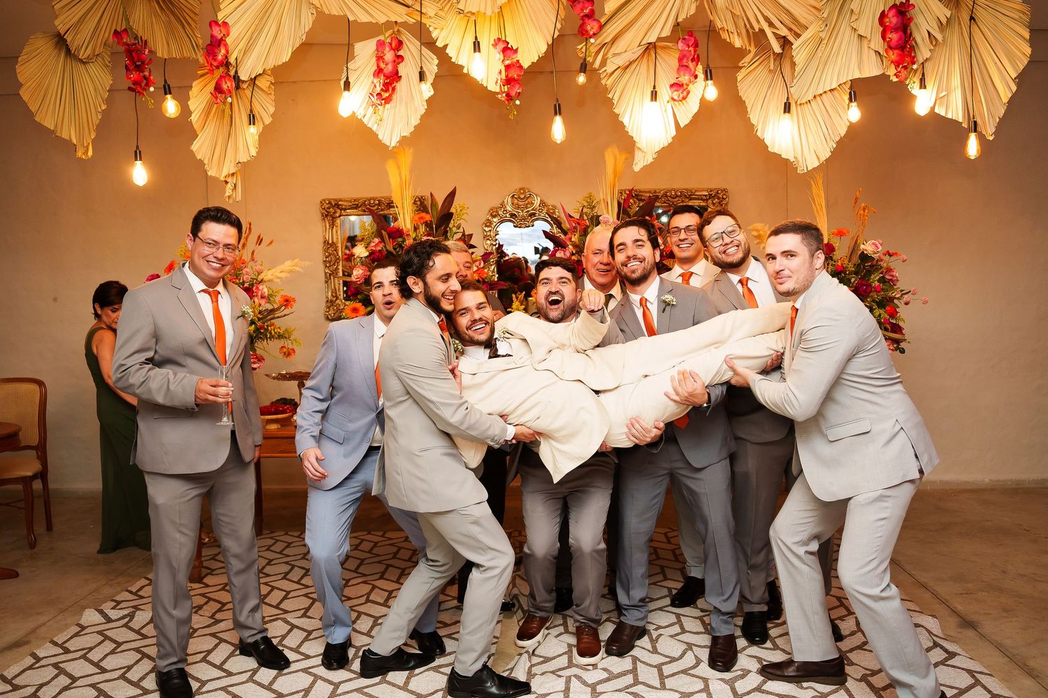 Groomsmen in gray suits lift the groom at a wedding celebration with tropical decor and warm string lights.