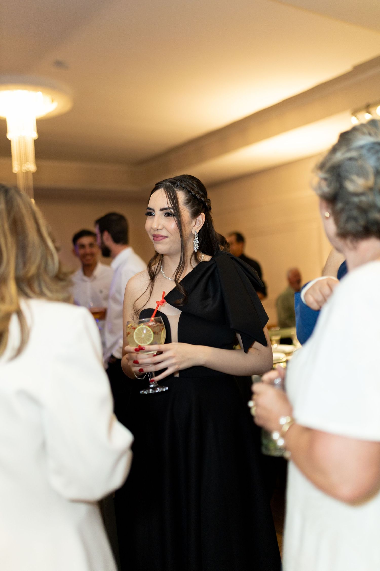 Woman in black dress holding a cocktail at an elegant indoor social event or formal party gathering.